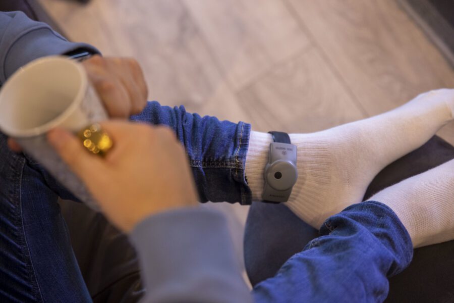 A close-up image of an ankle with an electronic device attached to it. The person is wearing casual clothes and holding a coffee mug.