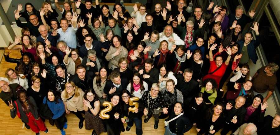 A large group of people standing together, looking up, smiling and waving at the camera. 