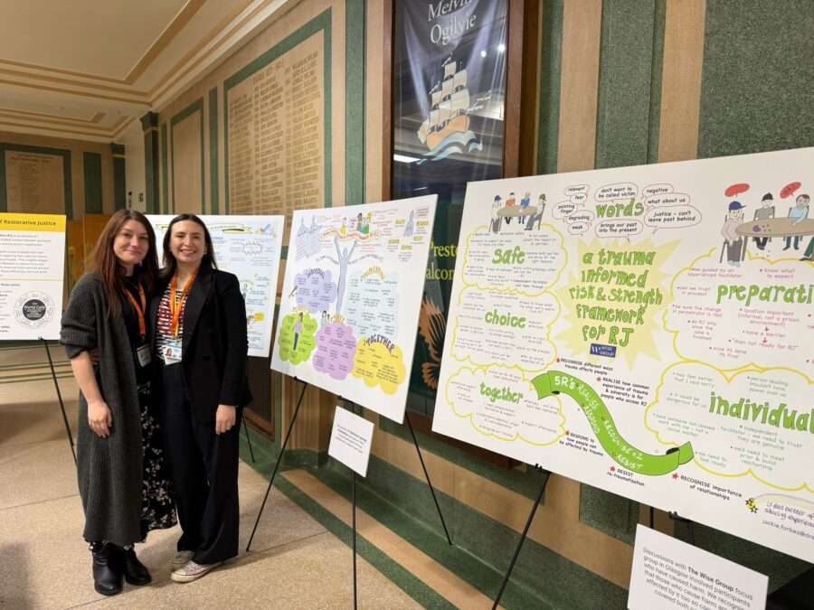 Two people stand beside a series of large illustrated boards displayed on easels in a hallway. The boards feature colourful graphics and text about restorative justice concepts, including trauma‑informed practice, safety, choice, and preparation. The setting appears to be an exhibition or learning event, with additional posters and signage visible along the wall behind them.