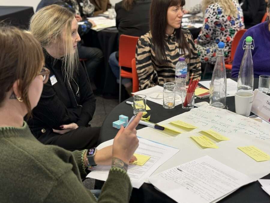 A group of people sit around a table during a workshop or group discussion. The table is covered with materials including papers, sticky notes, pens, water glasses, and bottles.