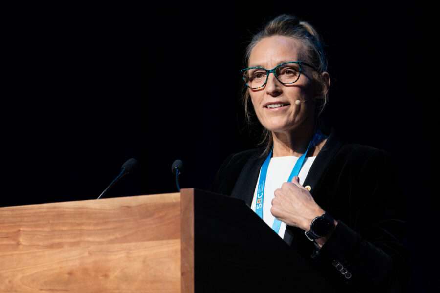 Tracey McFall making a speech at a podium, she is wearing glasses, a lanyard and a headset microphone