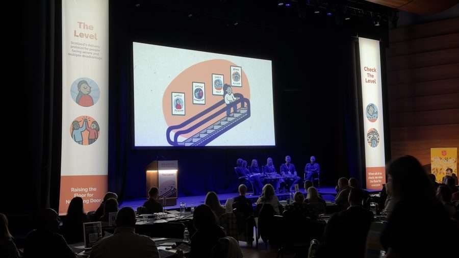 Six people on a dark stage. There is a large screen behind them and a long banner either side of the stage - they have illustrations on them that depict positive life outcomes.